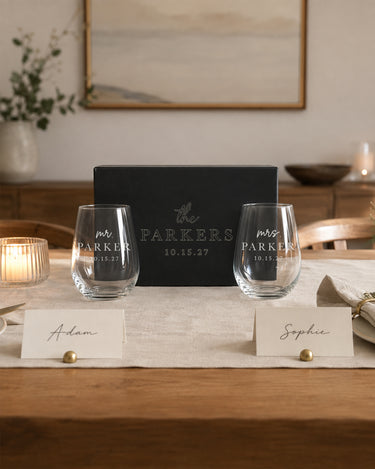 Two stemless wine glasses with lowercase script titles, bold surnames and dates beside a matching engraved black gift box.