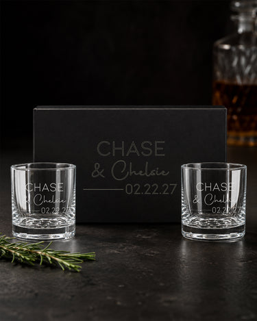 Two rocks glasses with print and script names, divider and date beside a matching engraved black magnetic gift box.
