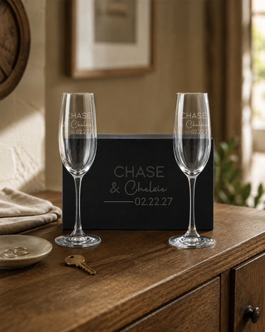 Champagne Flutes with two names in print and script, a divider and date beside a matching engraved black magnetic gift box.