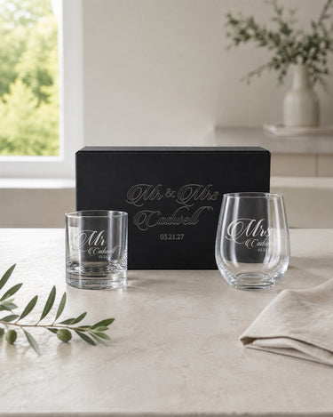 Mr rocks glass and Mrs stemless wine glass with a shared surname and date beside a matching engraved black magnetic gift box.