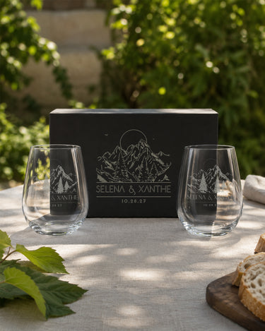 Stemless Wine Glasses engraved with mountain and evergreen artwork, names and a date beside a matching black magnetic gift box.