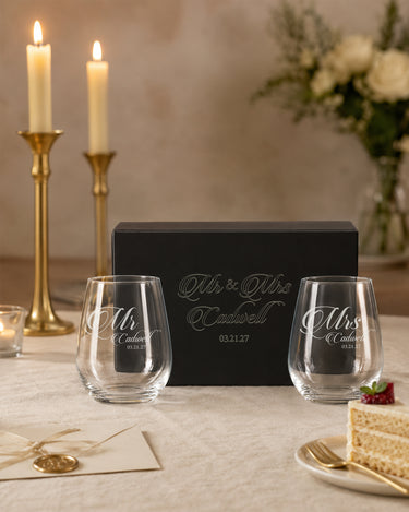 Two stemless wine glasses with Mr and Mrs script titles, a shared surname and date beside a matching engraved black gift box.