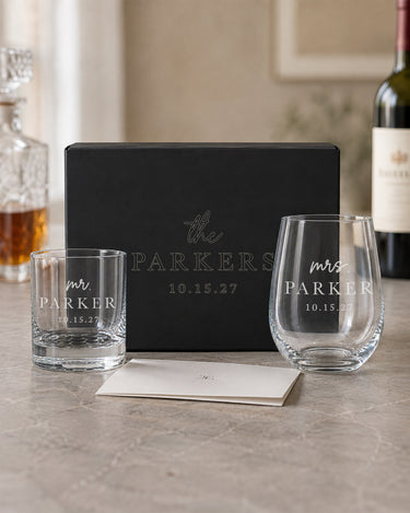 Rocks and stemless wine glasses with lowercase script titles, bold surnames and dates beside a matching engraved black gift box.