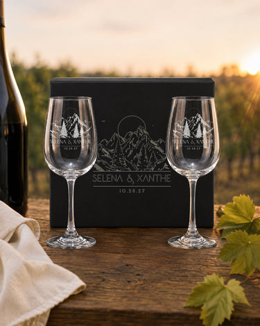Stemmed Wine Glasses engraved with mountain and evergreen artwork, names and a date beside a matching black magnetic gift box.