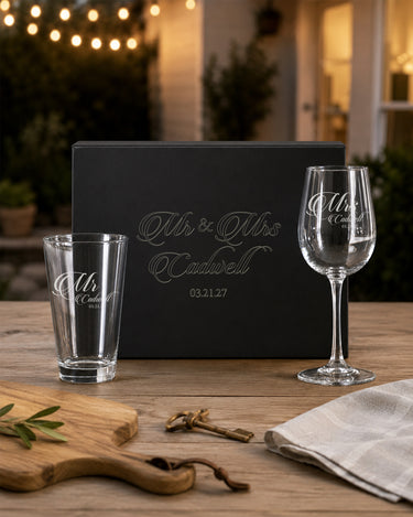 Mr pint glass and Mrs stemmed wine glass with a shared surname and date beside a matching engraved black magnetic gift box.