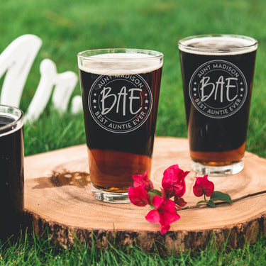 Two pint glasses on a table top. The glasses are etched with a design centered on the glass. The design has two outer circles, then inside it says "AUNT MADISON" on top rounded, and "BEST AUNTIE EVER" on the bottom rounded. Then there are 3 stars on each side, and another circle which inside of it says "BAE".