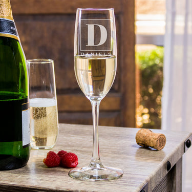 Champagne glass on a table. The glass has an etched design centered. The design is of a square with swirly horizontal lines, and in the middle is the letter "D" in caps. Below the square is the name "DANIELS" in all caps.