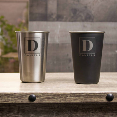 Two metal pints on a table. One pint is black with silver etching and the other pint is silver with black etching. The design is of a square with swirly horizontal lines, and in the middle is the letter "D" in caps. Below the square is the name "DANIELS" in all caps.