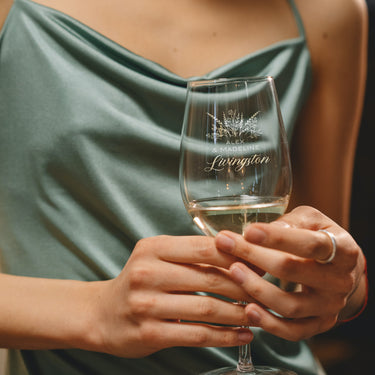Stemmed white wine glass on a table. The etched design is centered on the glass. The design is of a bouquet of flowers and beneath it is "Alex" and beneath that is "& Madeline" and beneath that is "Livingston". So, three lines of text: first name, send name and a last name. The first two names are in print font, the last name is in cursive.