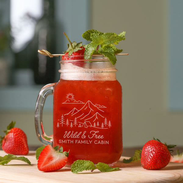Mason jar glass on a table. The glass has a design etched on the front and center. The design is outdoorsy, of a sun, clouds, mountains, trees and a cabin. Below the outdoors image is "Wild & Free" in cursive and below that is "SMITH FAMILY CABIN" in print font.