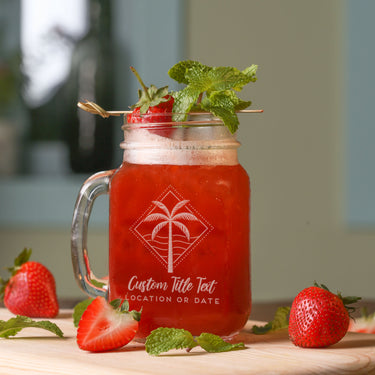 Mason jar glass on a table. The glass has a etched design centered on the front. The design etched on the glasses is a diamond shape line and dots, inside is a palm tree and waves. Below the design is "Custom Title Text" in cursive font, and below that is "LOCATION OR DATE" in printed font.