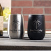 Two metal wine tumblers on a table. One tumbler is black and the etched design is in silver. The other tumbler is silver and the etched design is black. The design etched on the tumblers is diamond shape line and dots, inside is a palm tree and waves. Below the design is "Tina's Final Splash" in cursive font, and below that is "CABO, 2022" in printed font. The design is centered on the cup.