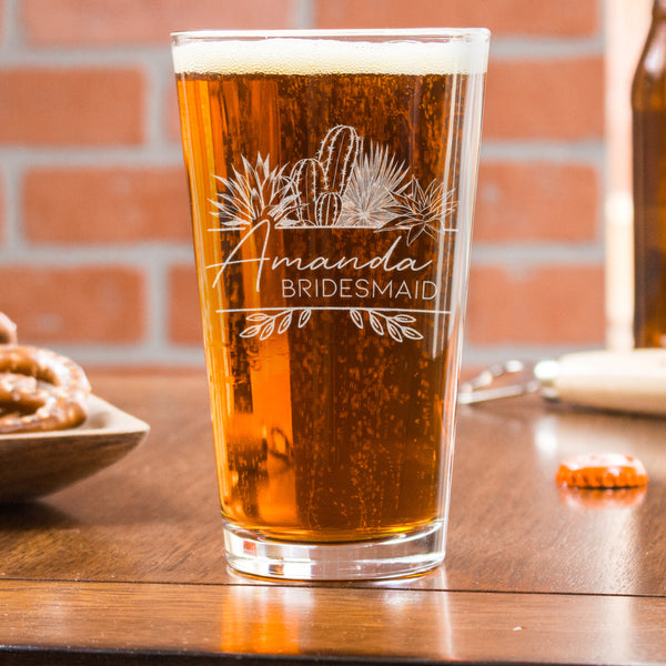Pint glass on a table. The glass has a design centered etched in the glass. The design is of different desert cactus plants and below is two lines, and between the lines it says the name "Amanda" in cursive and below the name is "BRIDESMAID" in all caps printed font.