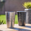 Black and silver metal pint cups on a table. The black pint has an etched design in silver. The silver pint has an etched design in black. The design is outdoorsy, of a sun, clouds, mountains, trees and a cabin. Below the outdoors image is "Wild & Free" in cursive and below that is "SMITH FAMILY CABIN" in print font.