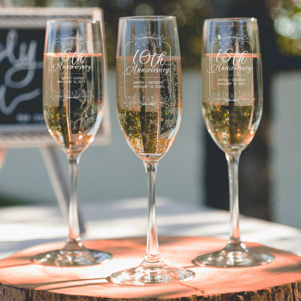 Stemmed champagne glasses with etched anniversary design. The design has a floral border with 5th, 10th, and 15th Anniversary inside the border along with two names for a couple and a wedding or anniversary date.