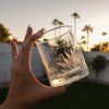 Whiskey rocks glass. The glass has a design etched on it that wraps around the entire glass. The design goes from the bottom of the glass all the way up past halfway. The design has a mix of desert cactus plants that wrap around, but on one side it says "Megan & Brian" in cursive and below that "PALM SPRINGS, CA" in printed font all caps.