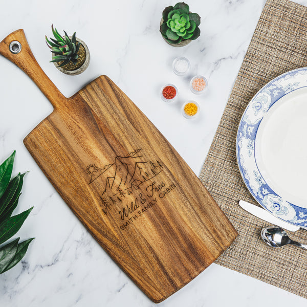 Long wood charcuterie board with handle on a table. The charcuterie board has an engraved design centered. The design is outdoorsy, of a sun, clouds, mountains, trees and a cabin. Below the outdoors image is "Wild & Free" in cursive and below that is "SMITH FAMILY CABIN" in print font.
