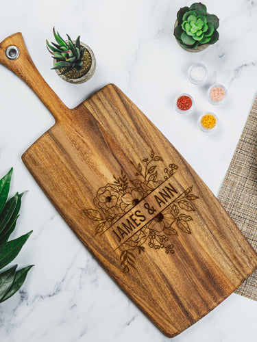 Birds eye view of a charcuterie board on a marble table top. The charcuterie board is wood, and is a long rectangular board with a handle. The board is engraved with a floral design and the names "JAMES & ANN" in the middle.