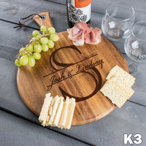 A charcuterie board with an engraved design in the center. The design has a large initial S with a section in the middle where it has a couple's names in a script font. A date is engraved to the top right of the names