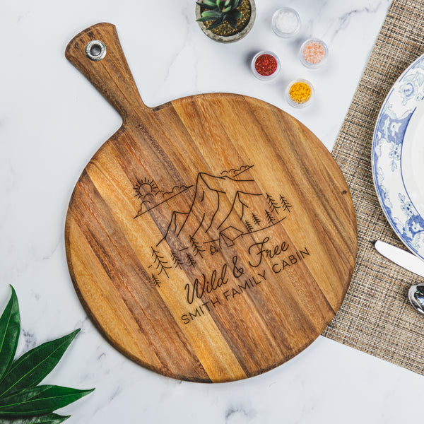 Round wood charcuterie board with handle on a table. The charcuterie board has an engraved design centered. The design is outdoorsy, of a sun, clouds, mountains, trees and a cabin. Below the outdoors image is "Wild & Free" in cursive and below that is "SMITH FAMILY CABIN" in print font.