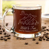 Glass coffee mug on a table. The glass has a design etched on the front and center. The design is outdoorsy, of a sun, clouds, mountains, trees and a cabin. Below the outdoors image is "Wild & Free" in cursive and below that is "SMITH FAMILY CABIN" in print font.