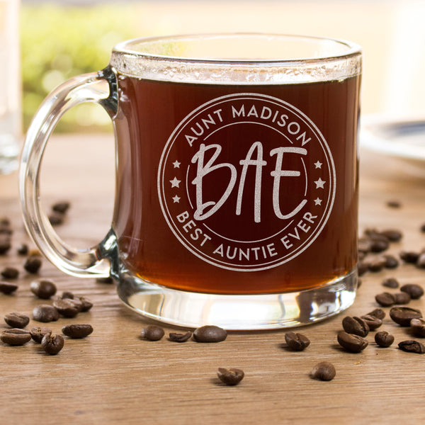 Glass mug filled with coffee on a table surrounded by coffee beans. The mug is etched with a design centered on the glass. The design has two outer circles, then inside it says "AUNT MADISON" on top, and "BEST AUNTIE EVER" on the bottom rounded. Then there are 3 stars on each side, and another circle which inside of it says "BAE".