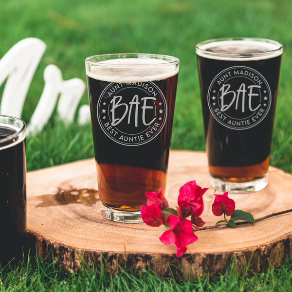 Two pint glasses on a table top. The glasses are etched with a design centered on the glass. The design has two outer circles, then inside it says "AUNT MADISON" on top rounded, and "BEST AUNTIE EVER" on the bottom rounded. Then there are 3 stars on each side, and another circle which inside of it says "BAE".