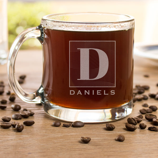 Glass coffee mug on a table. The glass has an etched design centered. The design is of a square with swirly horizontal lines, and in the middle is the letter "D" in caps. Below the square is the name "DANIELS" in all caps.