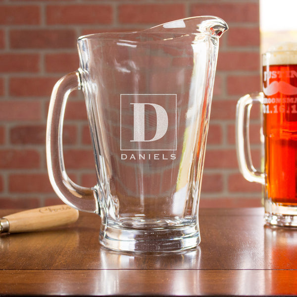 Glass pitcher on a table. The glass has an etched design centered. The design is of a square with swirly horizontal lines, and in the middle is the letter "D" in caps. Below the square is the name "DANIELS" in all caps.
