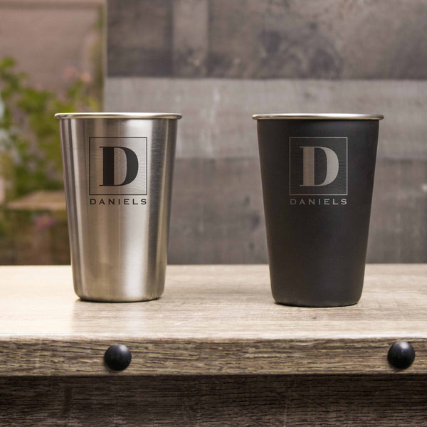 Two metal pints on a table. One pint is black with silver etching and the other pint is silver with black etching. The design is of a square with swirly horizontal lines, and in the middle is the letter "D" in caps. Below the square is the name "DANIELS" in all caps.