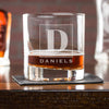 Whiskey glass on a table. The glass has an etched design centered. The design is of a square with swirly horizontal lines, and in the middle is the letter "D" in caps. Below the square is the name "DANIELS" in all caps.