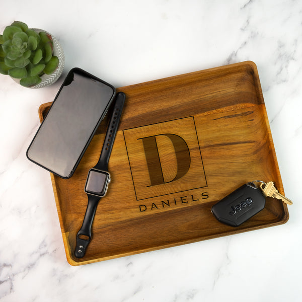 Wooden tray on a marble table top. The wooden tray has an engraved design centered in the middle. The design is of a square with swirly horizontal lines, and in the middle is the letter "D" in caps. Below the square is the name "DANIELS" in all caps.