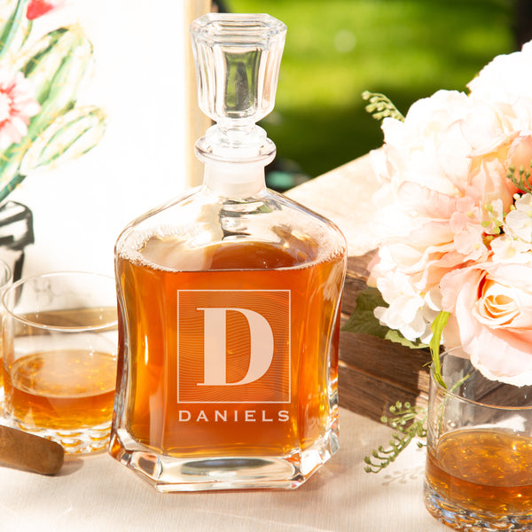 Glass whiskey decanter on a table. The glass has an etched design centered. The design is of a square with swirly horizontal lines, and in the middle is the letter "D" in caps. Below the square is the name "DANIELS" in all caps.