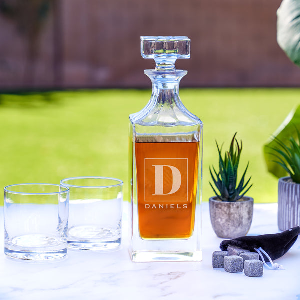 Tall glass whiskey decanter on a table. The glass has an etched design centered and in the middle. The design is of a square with swirly horizontal lines, and in the middle is the letter "D" in caps. Below the square is the name "DANIELS" in all caps.