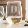 Stemless white wine glass on a table. The glass has an etched design centered. The design is of a square with swirly horizontal lines, and in the middle is the letter "D" in caps. Below the square is the name "DANIELS" in all caps.