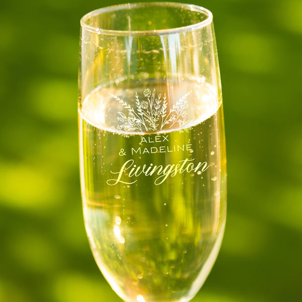 Champagne toasting glass on a table. The etched design is centered on the glass. The design is of a bouquet of flowers and beneath it is "Alex" and beneath that is "& Madeline" and beneath that is "Livingston". So, three lines of text: first name, send name and a last name.