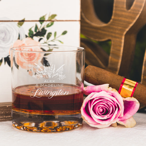 Etched whiskey glass on a table. The etched design is centered on the glass. The design is of a bouquet of flowers and beneath it is "Alex" and beneath that is "& Madeline" and beneath that is "Livingston". So, three lines of text: first name, send name and a last name.