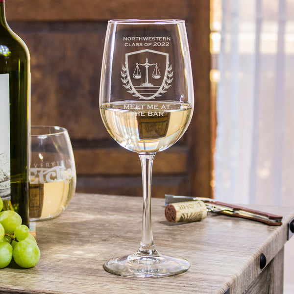 Stemmed white wine glass on a table. The glass has an etched design centered. The design has "NORTHWESTERN" in all caps, and centered below says "CLASS OF 2022". Under the text is an image of a scale, inside of a shield with wreaths to represent the justice system. Below the design is the words "MEET ME AT THE BAR" in all caps centered. This design is a play on words of the bar exam and a drinking bar.