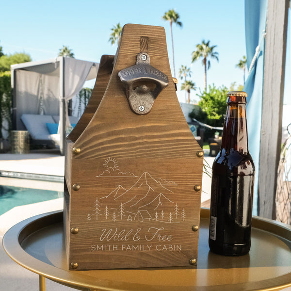 Wooden beer caddy on a table. The caddy has an engraved design on one side centered. The design is outdoorsy, of a sun, clouds, mountains, trees and a cabin. Below the outdoors image is "Wild & Free" in cursive and below that is "SMITH FAMILY CABIN" in print font.
