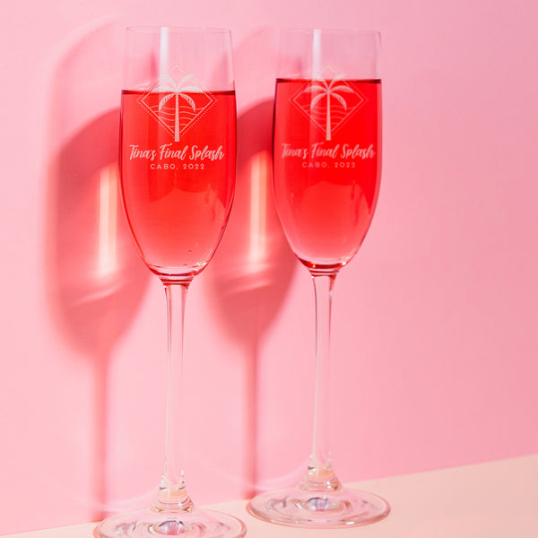 Two stemmed champagne glasses on a table. The champagne glasses have a etched design centered on the front. The design etched on the glasses is a diamond shape line and dots, inside is a palm tree and waves. Below the design is "Tina's Final Splash" in cursive font, and below that is "CABO, 2022" in printed font.