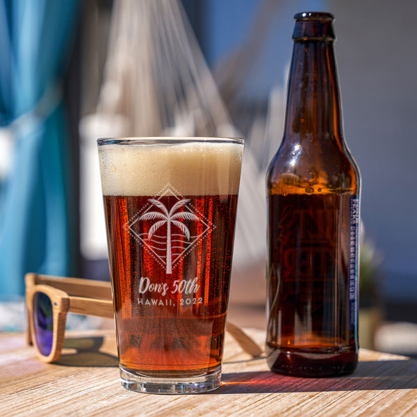Pint glass on a table. The glass has a etched design centered on the front. The design etched on the glasses is a diamond shape line and dots, inside is a palm tree and waves. Below the design is "Don's 50th" in cursive font, and below that is "HAWAII, 2022" in printed font.