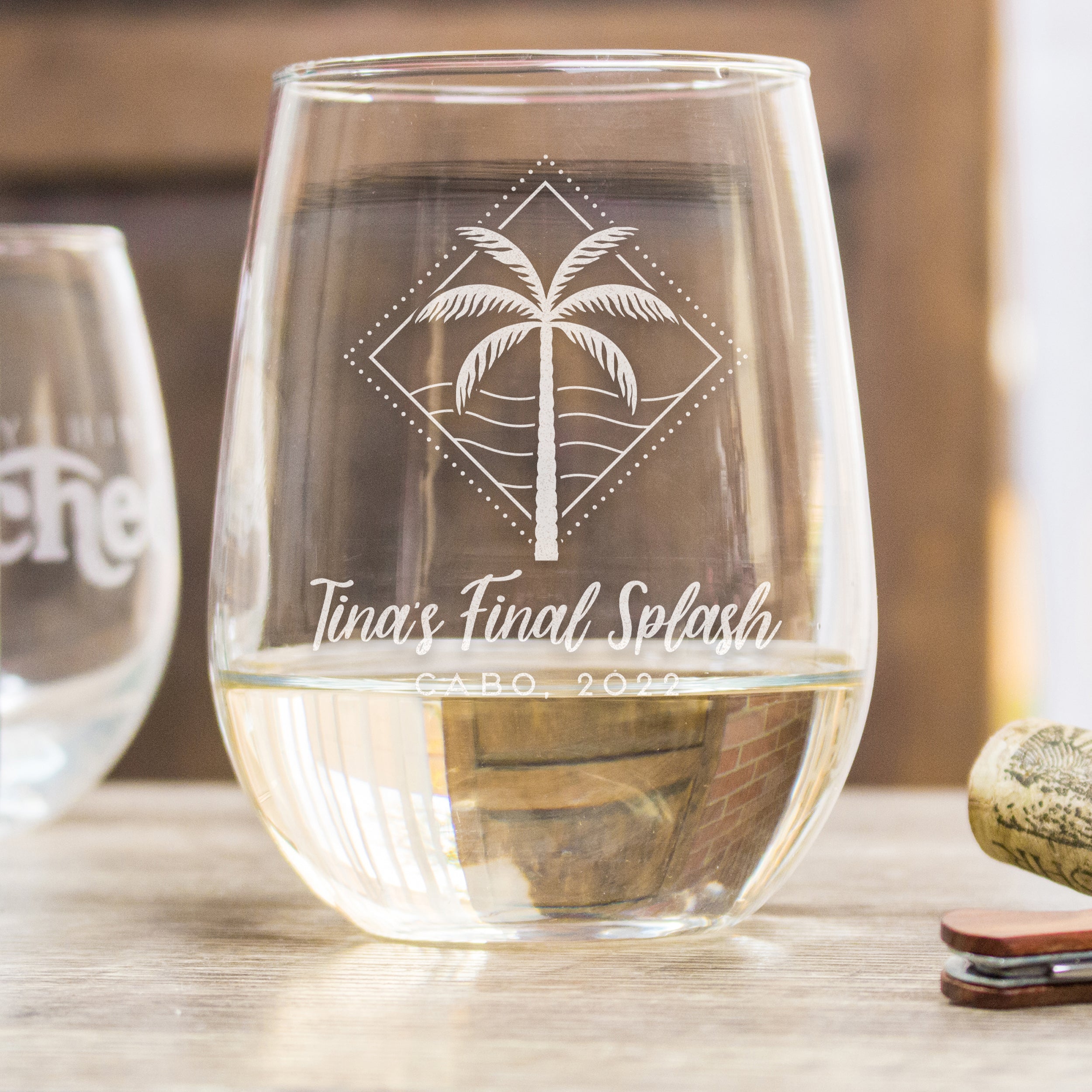 Stemless white wine glass on a table. The glass has a etched design centered on the front. The design etched on the glasses is a diamond shape line and dots, inside is a palm tree and waves. Below the design is "Custom Title Text" in cursive font, and below that is "LOCATION OR DATE" in printed font. 