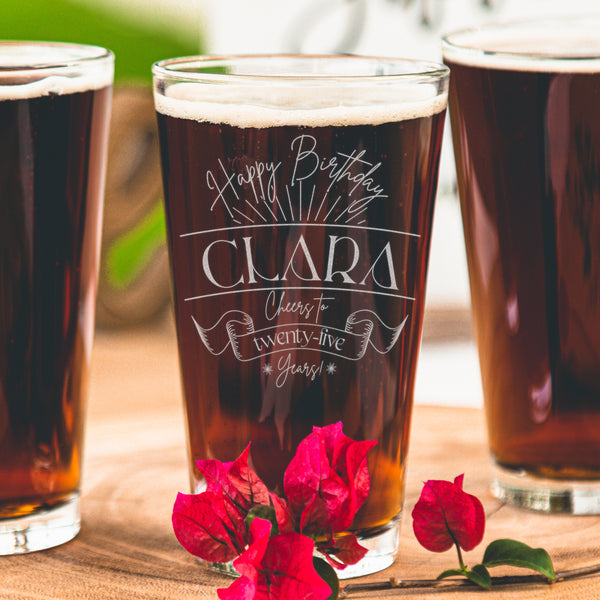 Pint glass on a table. The glass has an etched design centered on the front. The design has "Happy Birthday" in cursive rounded at the top. Below is some firework lines. Then is the name "CLARA" in between two horiztonal lines in printed all caps font. Then it says "Cheers to" in cursive, then a banner that says "twenty-five" inside, and "years!" at the bottom in cursive. The entire design is centered on top of one another.
