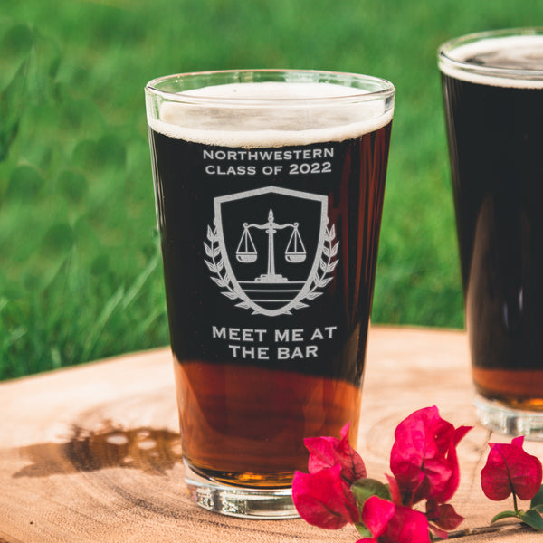 Pint glass on a table. The glass has an etched design centered. The design has "NORTHWESTERN" in all caps, and centered below says "CLASS OF 2022". Under the text is an image of a scale, inside of a shield with wreaths to represent the justice system. Below the design is the words "MEET ME AT THE BAR" in all caps centered. This design is a play on words of the bar exam and a drinking bar.