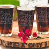 Pint glass on a table. The glass has a design etched on the front and center. The design is outdoorsy, of a sun, clouds, mountains, trees and a cabin. Below the outdoors image is "Wild & Free" in cursive and below that is "SMITH FAMILY CABIN" in print font.