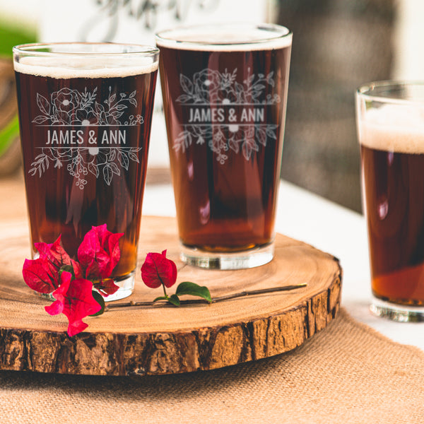 Two pint glasses on a marble table top. The glasses are etched with a floral design and in the middle of the design is a rectangle shape cutting off the flowers and has the names "JAMES & ANN". The etched design is placed center on the glass.