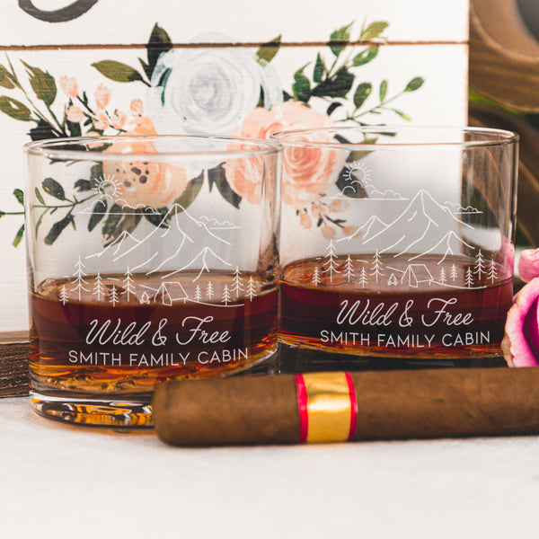 Whiskey glass on a table. The glass has an etched design centered. The design is outdoorsy, of a sun, clouds, mountains, trees and a cabin. Below the outdoors image is "Wild & Free" in cursive and below that is "SMITH FAMILY CABIN" in print font.
