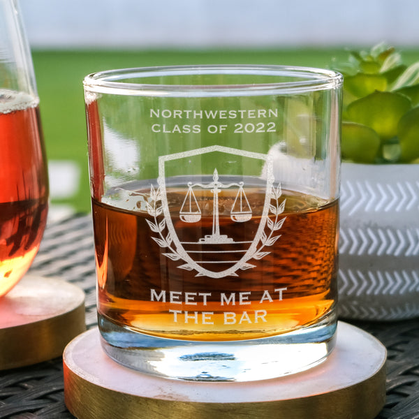 Whiskey glass on a table. The glass has an etched design centered. The design has "NORTHWESTERN" in all caps, and centered below says "CLASS OF 2022". Under the text is an image of a scale, inside of a shield with wreaths to represent the justice system. Below the design is the words "MEET ME AT THE BAR" in all caps centered. This design is a play on words of the bar exam and a drinking bar.