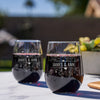 Two stemless red wine glasses on a marble table top. The glasses are etched with a floral design and in the middle of the design is a rectangle cutting off the flowers and has the names "JAMES & ANN". The etched design is placed center on the glass.