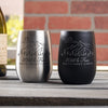 Black and silver metal wine tumblers on a table. The black tumbler has an etched design in silver. The silver tumbler has an etched design in black. The design is outdoorsy, of a sun, clouds, mountains, trees and a cabin. Below the outdoors image is "Wild & Free" in cursive and below that is "SMITH FAMILY CABIN" in print font.